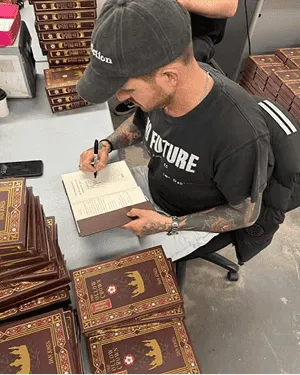 A person wearing a black cap and t-shirt is seated at a table, signing a book. Surrounding them are stacks of books with ornate brown covers featuring a crown and red emblem.