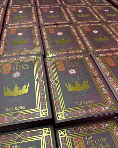 Stacks of "The Hollow Crown" books by Dan Jones are neatly arranged, featuring ornate covers with a gold crown design and decorative floral borders.